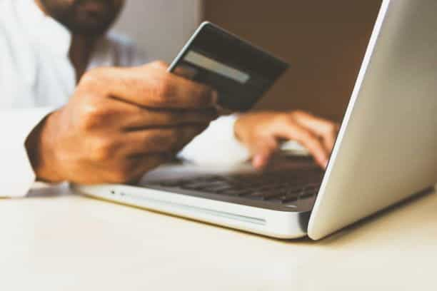 A dark-skinned man in a button down shirt holds a credit card next to his computer as he types.