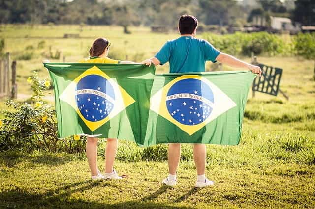 A man and a woman turn their backs to the camera and hold Brazilian flags behind them.