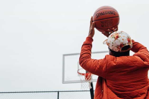 The backside of a man in an orange jacket and baseball cap throwing a basketball at a basketball hoop outside.