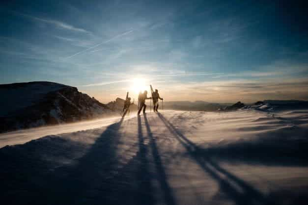 Three people with skiing gear walk towards the summit of a snow covered mountain as the sun sets.