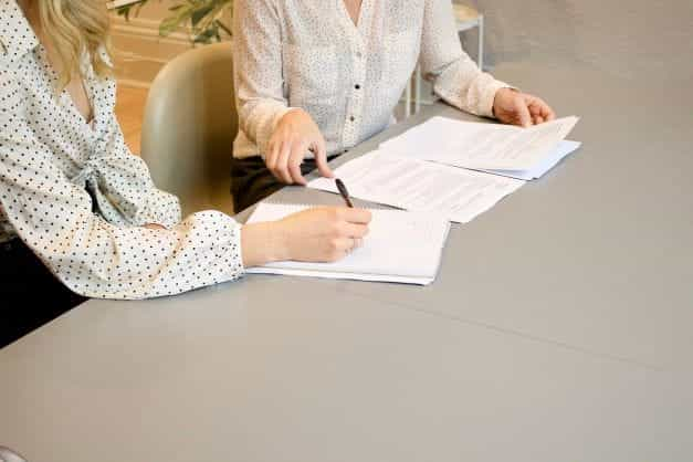 Two women sitting at a table with documents with one of them signing on them.