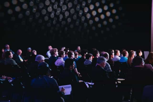 A group of people sit in the audience of a conference, watching a presentation.