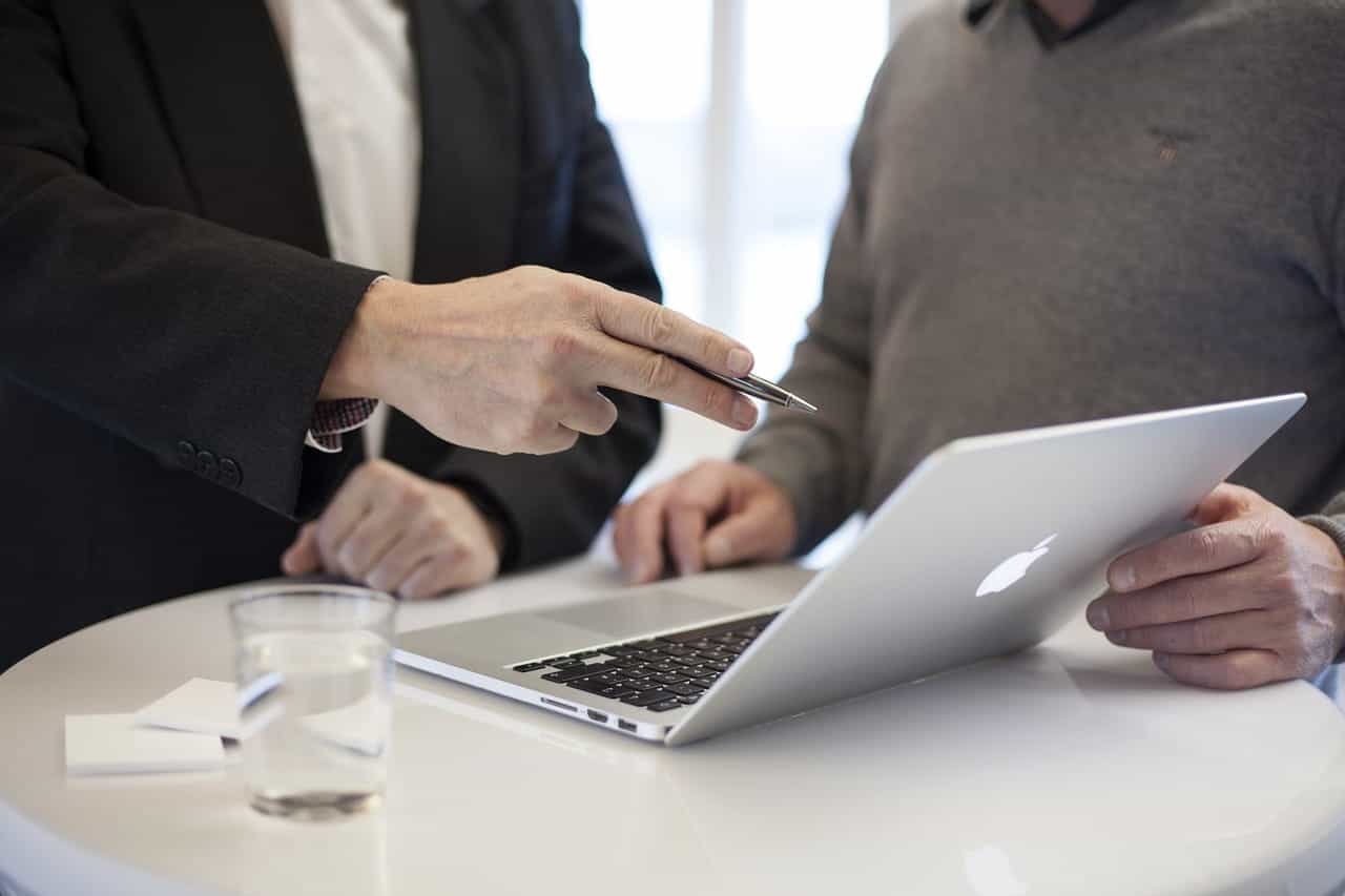 A person standing and pointing at an open laptop which is sitting on a desk next to another person standing and observing the laptop screen.