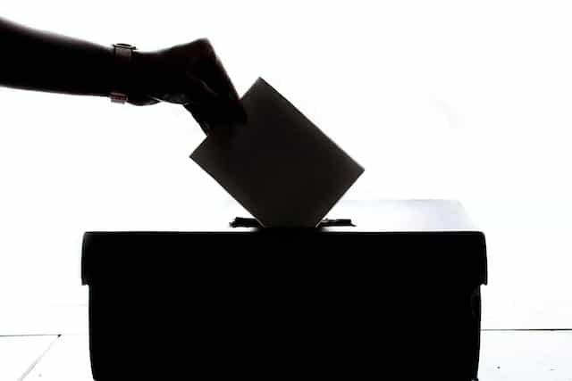 A silhouette of a person’s hand slipping an envelope into a ballot box during an election.