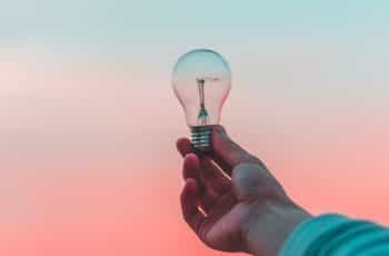 A person’s outstretched hand holding up a lightbulb against a blue and orange background.