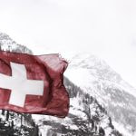 A red flag with a white cross against a snowy mountain range.